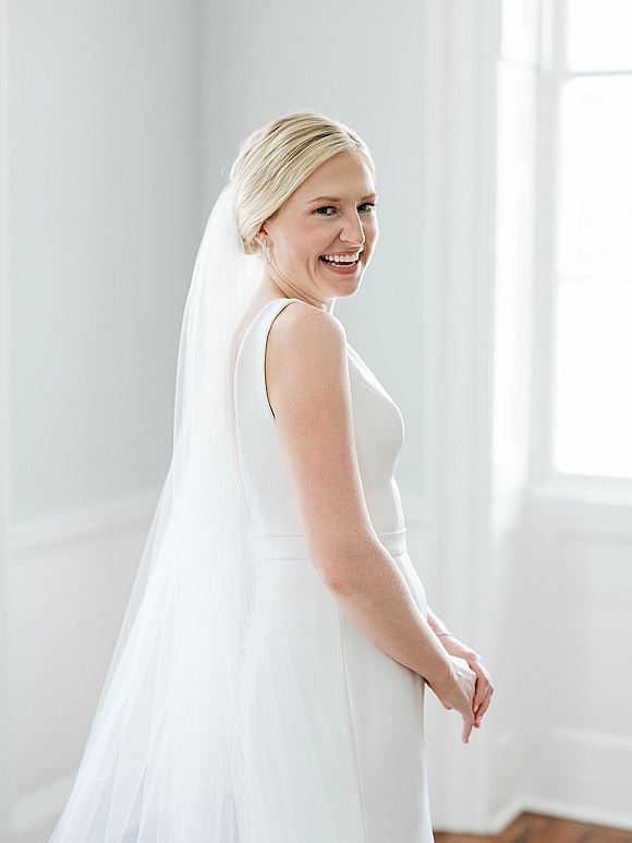 Bridal portrait of a bride in a simple wedding dress with a long veil, smiling in side profile by a large window in natural light