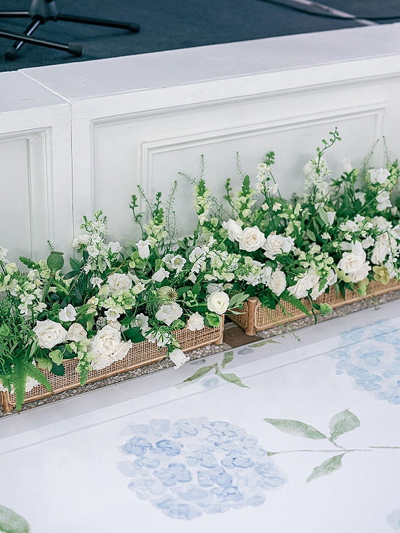 Wedding floral arrangements in wicker baskets with white flowers and greenery lining a stage platform near a microphone stand