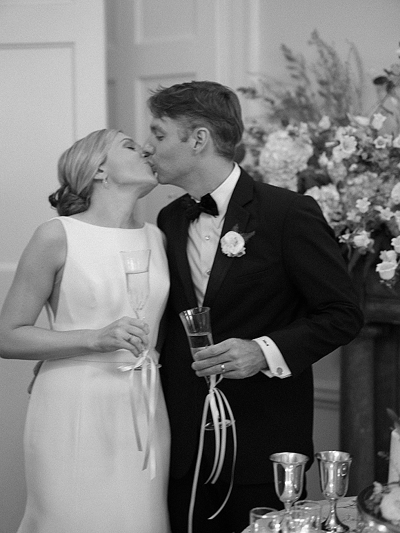Wedding kiss as bride and groom sip champagne flutes with ribbons at an indoor reception table setting, tuxedo and sleeveless dress close up