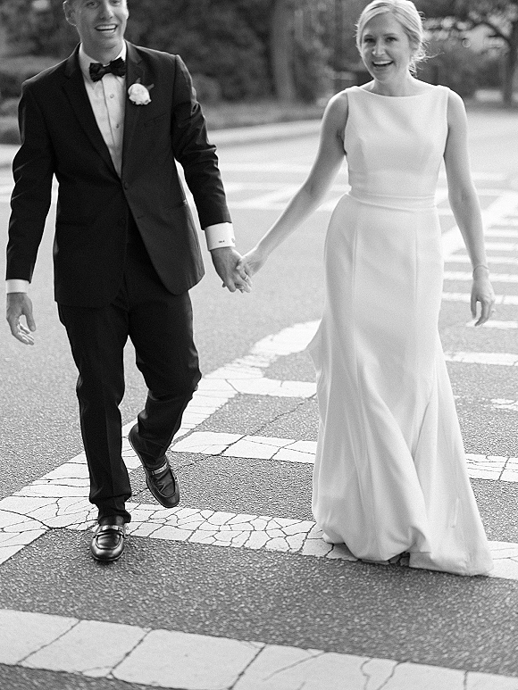 Wedding couple portrait of bride and groom holding hands, laughing as they walk across a crosswalk in tuxedo and sleeveless gown with belt