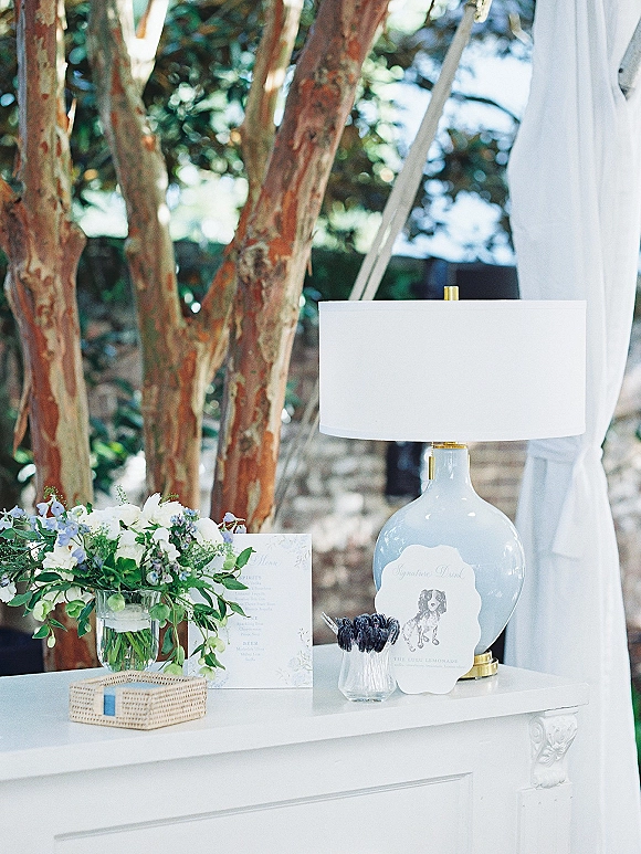 Welcome table decor with a wedding welcome table display, lamp, blue-and-white florals, menus, stirrers, and signage by greenery and stone wall