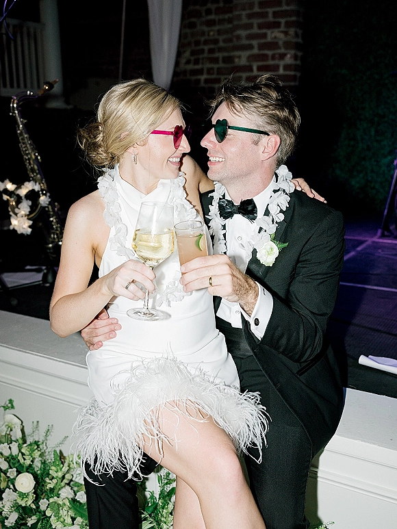 Wedding reception couple in heart sunglasses raising a newlywed toast, bride in feather-trim dress on groom’s lap before a brick wall stage