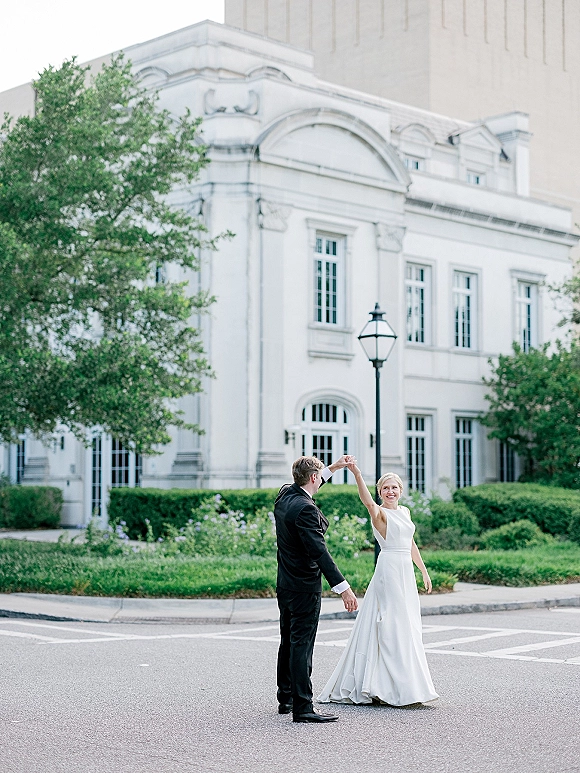 Couple portrait of bride in a wedding dress twirling with groom in tuxedo, holding hands on a crosswalk by a white arched building