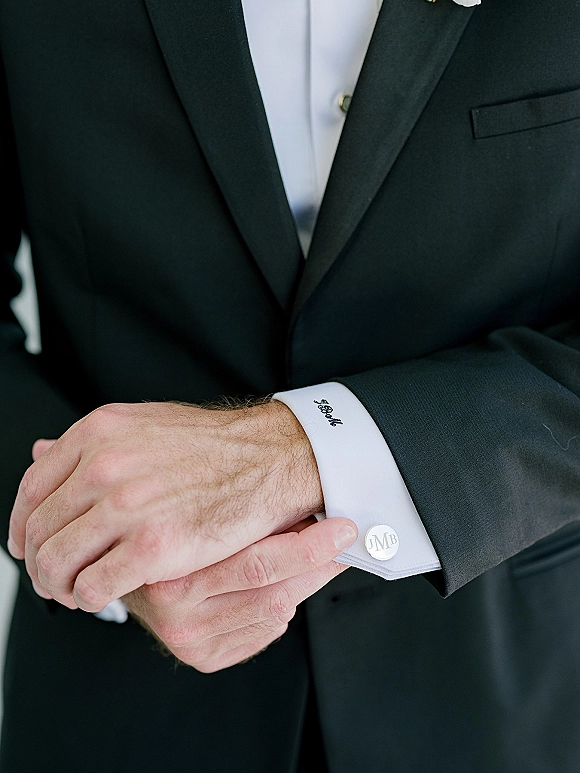 Groom accessories in close-up, hands adjusting wedding cufflinks on a white dress shirt under a black tuxedo jacket, neutral blur background