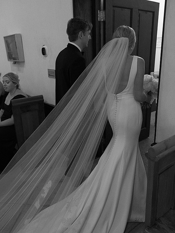 Wedding processional with bride walking down aisle in satin gown and long veil, holding bouquet, as groom in tux waits by church doorway