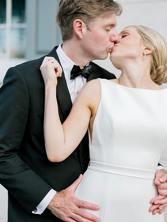 Wedding kiss portrait of bride and groom kissing, groom in black tuxedo with satin bow tie, bride in high-neck gown by white wall window