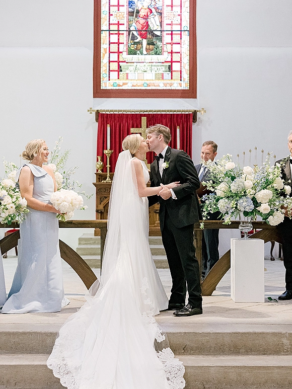 Wedding kiss as the bride and groom share their first kiss ceremony at the church altar, her cathedral veil trailing beneath stained glass.