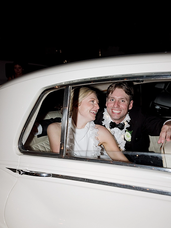Wedding getaway car with bride and groom smiling in a vintage white car, her gown and floral leis visible on a dark street at night