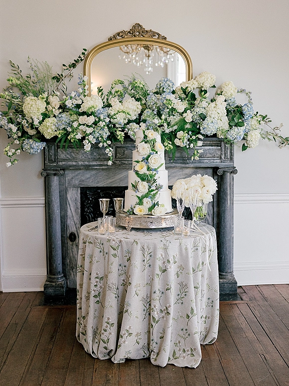 Wedding cake table with a tiered wedding cake topped with roses and hydrangeas, candles and champagne flutes before a vintage mirror mantel