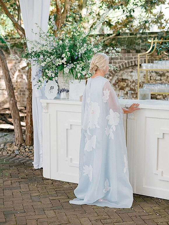 Wedding bar setup with an outdoor wedding bar, white draping, lush white flowers and greenery, glassware, and menu sign on a brick patio