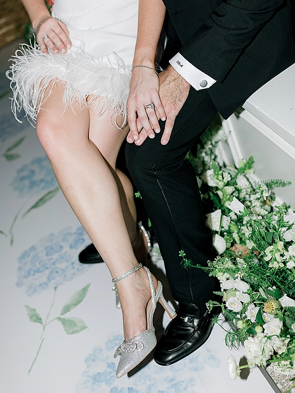 Wedding rings close-up on bride and groom hands, diamond engagement ring and band stack against tuxedo cufflinks and fringe dress accents
