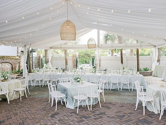 Reception tablescape under a tent wedding reception with blue patterned linens, candles and small florals, wicker pendants, and string lights in a courtyard