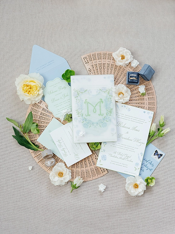 Invitation suite flat lay with monogram card, reply card, rings, ring box, and yellow rose accents on a linen backdrop