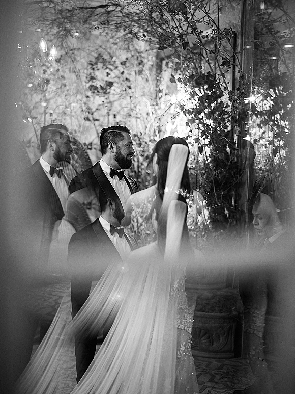 Ceremony moment as groom in black tuxedo watches bride in lace gown and long veil at an indoor venue with hanging lights