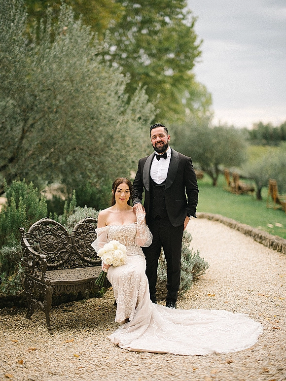 Couple portrait of bride in strapless lace dress holding a white rose bouquet as groom in black tuxedo stands by ornate bench in garden