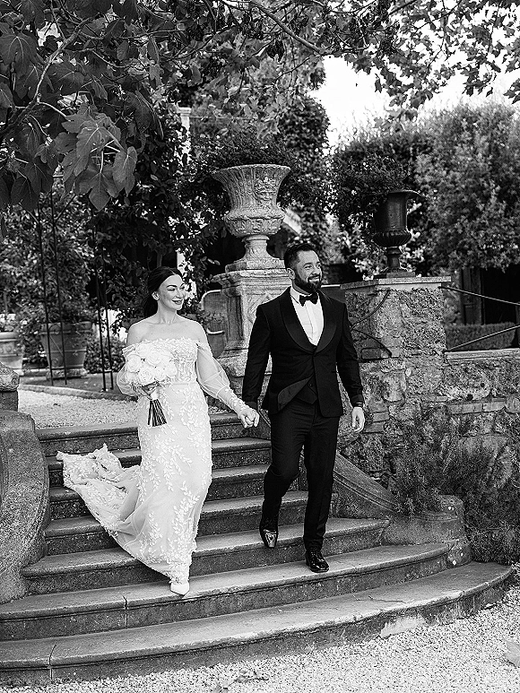 Wedding couple portrait of bride in veil holding bouquet and groom in tuxedo walking hand in hand by a garden stone staircase