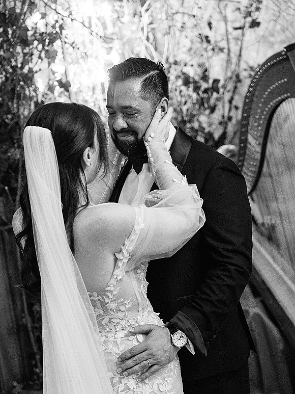 Couple portrait in a black and white wedding portrait as the bride in a lace veil cups the groom’s face, eyes closed, amid garden greenery