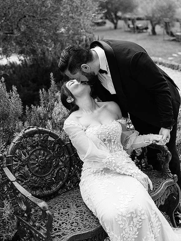 Wedding kiss portrait of bride and groom kissing on an ornate metal bench, her off-the-shoulder lace dress and his tuxedo in garden greenery