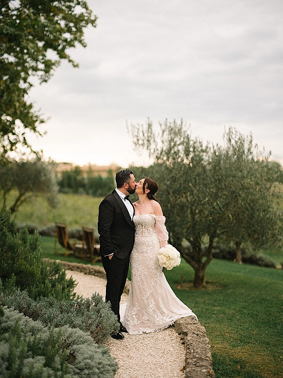 Wedding kiss portrait of bride and groom kissing on a gravel garden path, her strapless lace gown with tulle sleeves and white rose bouquet