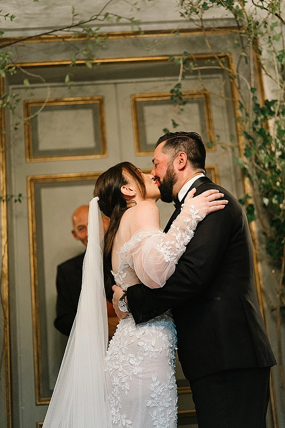 Wedding kiss portrait of bride and groom kissing in an intimate ceremony kiss moment, her long veil flowing before paneled doors with gold trim