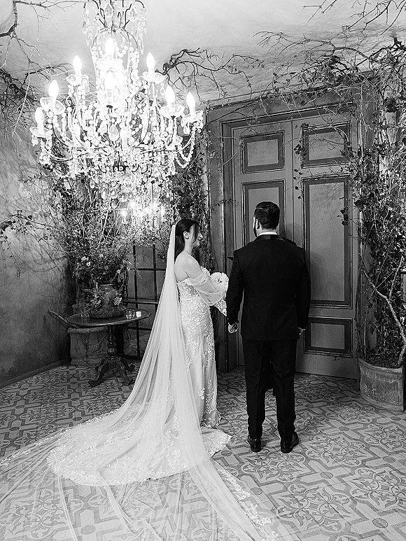 Couple portrait in black and white, bride and groom holding hands from behind beneath chandelier, candles, and lush greenery indoors