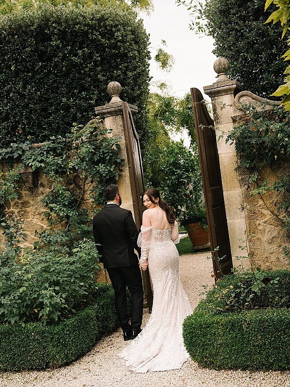 Couple portrait of bride and groom holding hands walking through an open wooden garden gate, her lace gown train trailing on gravel path