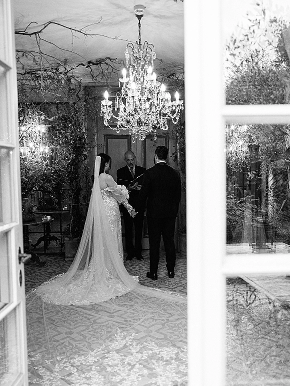 Wedding ceremony with couple exchanging vows before an officiant, bride in lace dress and veil holding bouquet under chandelier and candles