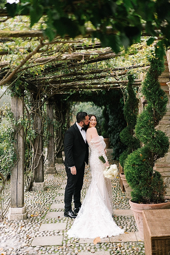 Couple portrait of groom kissing bride’s cheek as she looks over her shoulder, holding a bouquet under a vine-covered pergola walkway