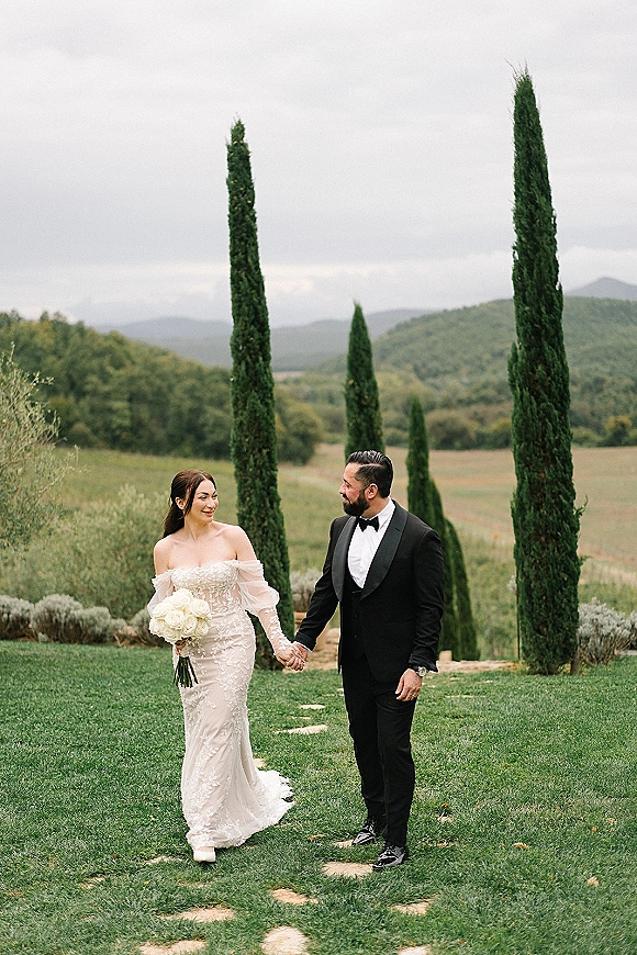 Couple portrait of bride and groom holding hands on a green lawn, bride with white rose bouquet, groom in tuxedo under cloudy hills