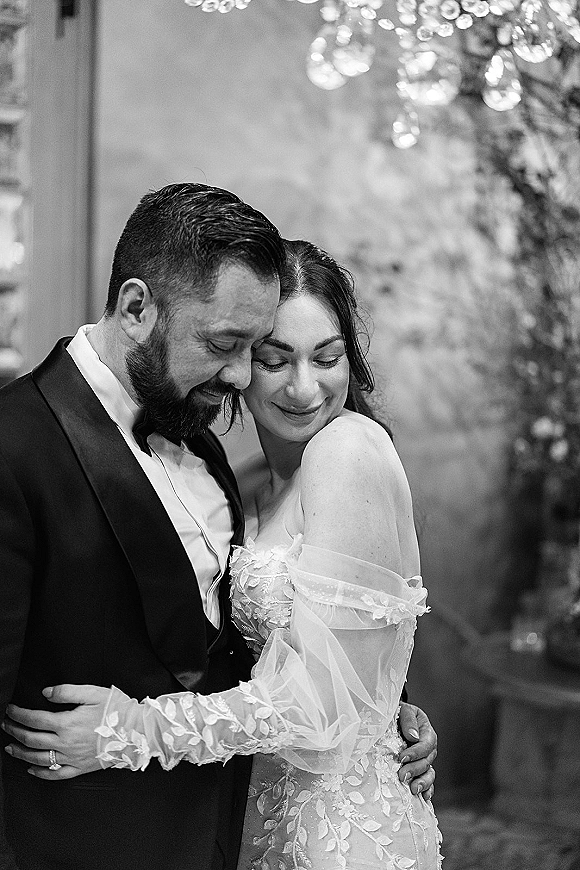 Couple portrait in a black and white wedding portrait as bride and groom embrace with a forehead touch under hanging lights and greenery