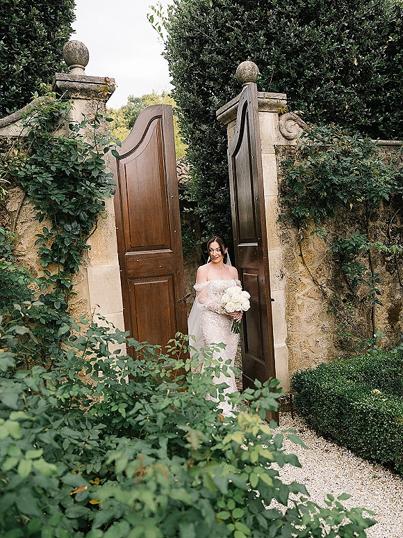 Bridal portrait of a bride holding bouquet in a strapless lace gown and cathedral veil, standing by ivy stone wall and wooden gates