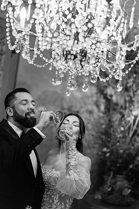 Wedding toast moment as newlyweds toasting with champagne flutes, bride in lace sleeves and groom in tuxedo under a chandelier