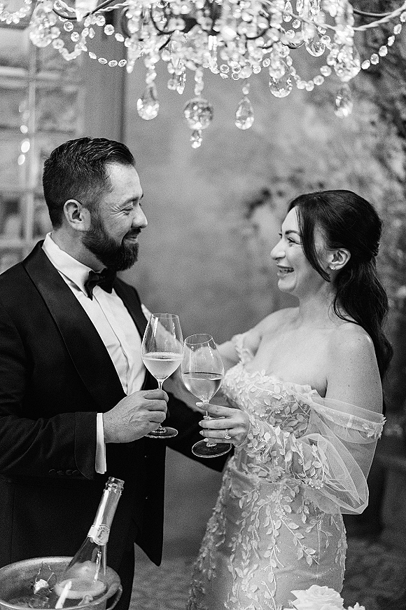 Wedding toast as bride in off-the-shoulder gown and groom in black tuxedo clink wine glasses beneath a crystal chandelier indoors