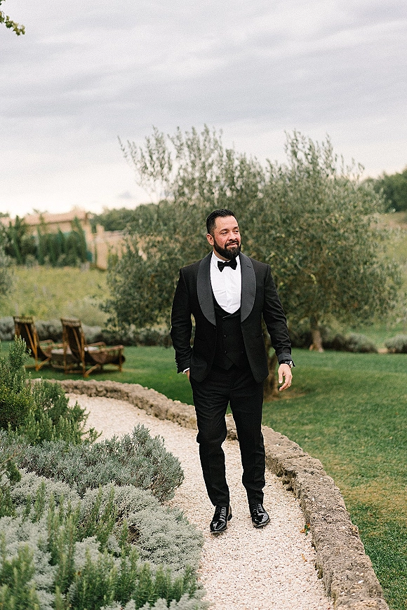 Groom portrait in a black tuxedo groom look, walking a gravel garden path under a cloudy sky with lounge chairs behind