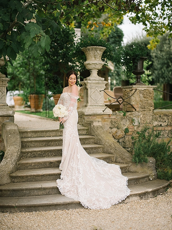 Bridal portrait of a bride holding bouquet of white roses in an off-the-shoulder lace gown with long train on a stone staircase in a garden