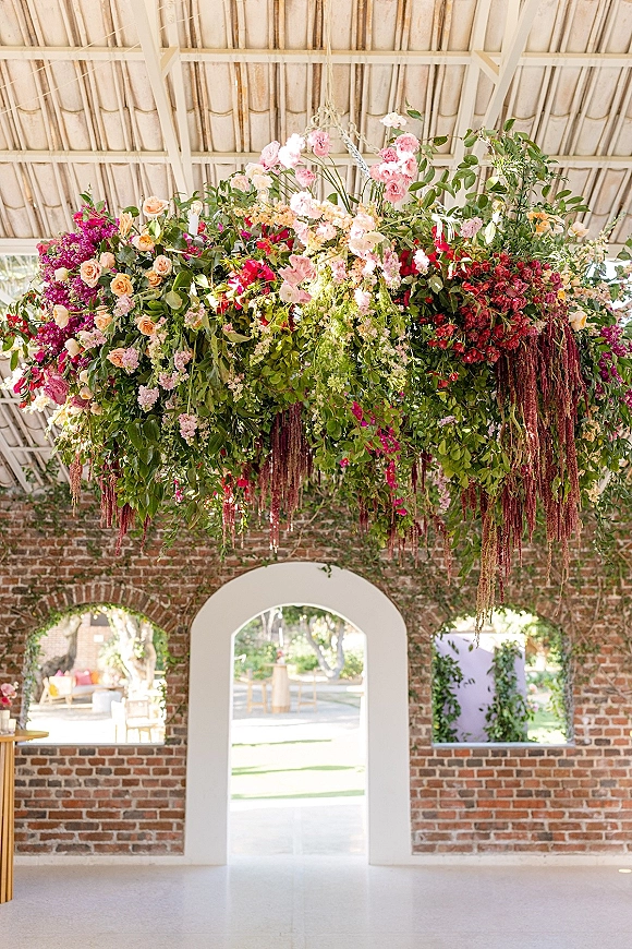 Hanging floral installation wedding floral chandelier with pink and peach roses and cascading amaranthus under white beams by brick arches