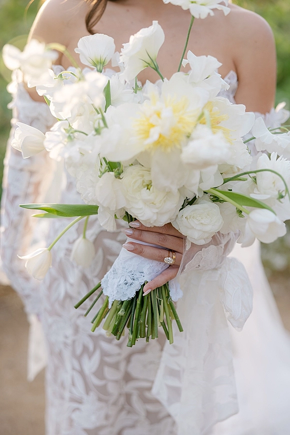 Bridal bouquet, white wedding bouquet cradled in lace-sleeved hands with engagement ring and nude nails against sunlit greenery