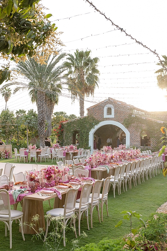 Reception tablescape at an outdoor wedding reception with long banquet tables, pink florals and candles under string lights in a brick courtyard