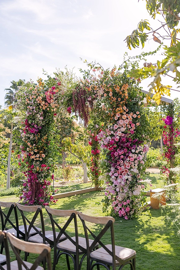 Floral ceremony arch with pink roses and hanging florals, framed by lush greenery garland and wooden chairs on a garden lawn