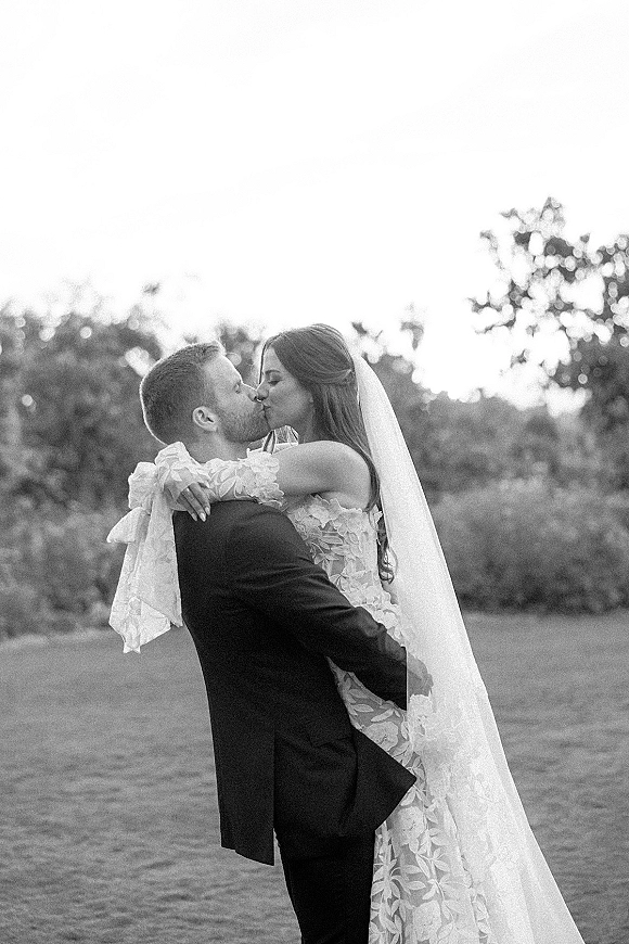 Wedding kiss portrait of bride and groom kissing as he lifts her, veil blowing over lace long-sleeve gown on a tree-lined lawn