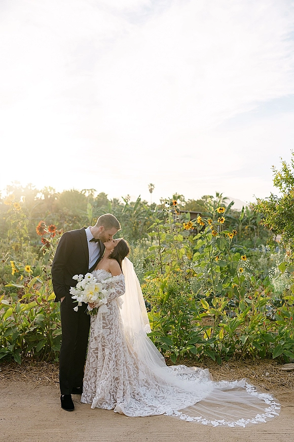 Wedding kiss portrait of bride and groom kissing at golden hour, her lace gown and veil flowing as she holds a white bouquet in a wildflower garden