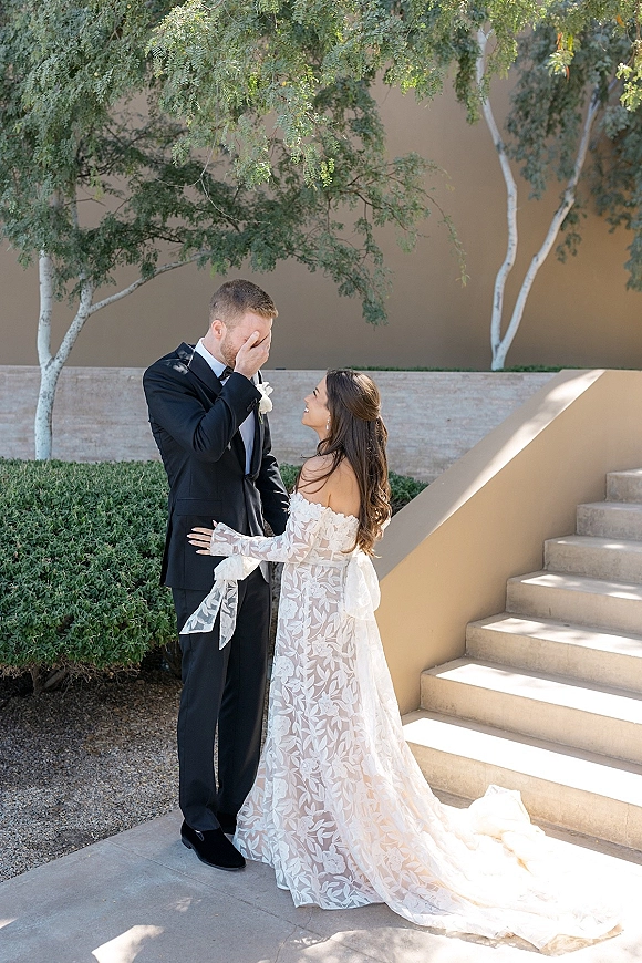 First look moment as bride in off-the-shoulder lace gown reaches groom in black tuxedo covering his face on a sunlit staircase walkway