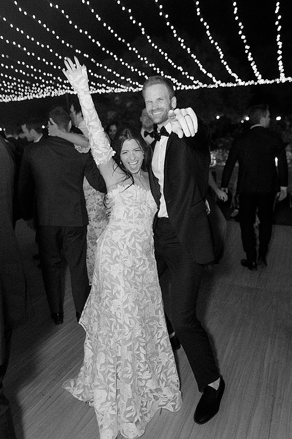 Wedding dance moment as bride in lace gown and gloves twirls with groom in tuxedo under string lights, guests cheering on the dance floor at night