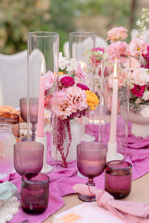 Reception tablescape with a pink table runner, taper candles in glass hurricane holders, colorful floral centerpieces, and goblets amid outdoor greenery