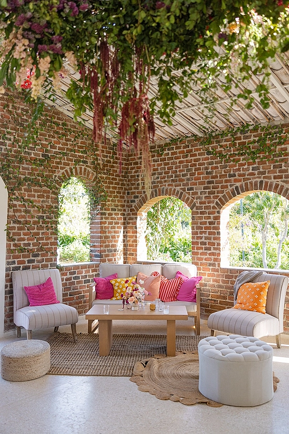 Wedding lounge decor with sofa seating, pillows, and candlelit coffee table topped with floral centerpiece beneath hanging greenery in a brick, arched-window venue