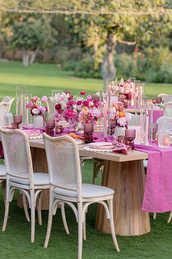 Reception tablescape with outdoor reception table set with pink runner, bright florals, taper candles, and rattan chairs under string lights on a lawn