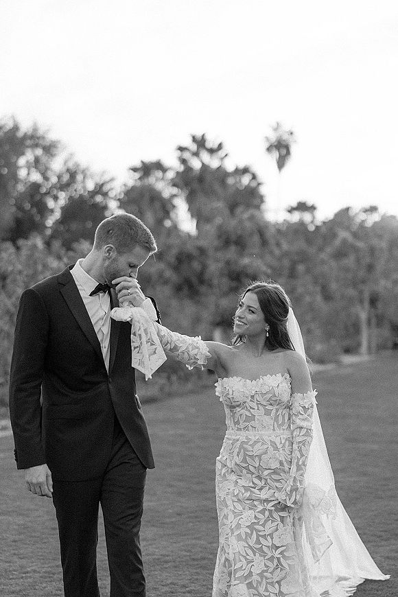 Couple portrait in a black and white wedding portrait style, groom kissing bride’s hand as her veil flows on a lawn with palm trees