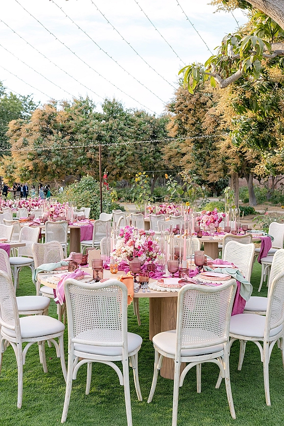Reception tablescape at an outdoor wedding reception with pink linens, purple goblets, floral centerpieces, and glowing candles under string lights on a lawn
