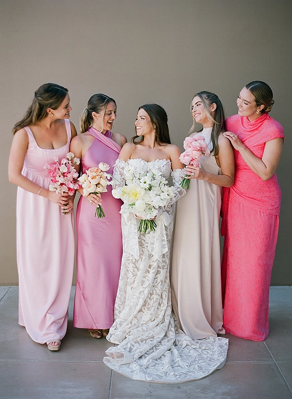 Bride with bridesmaids in pink dresses laughing together, holding bouquets beside a neutral wall, bride in off-the-shoulder lace gown and updo
