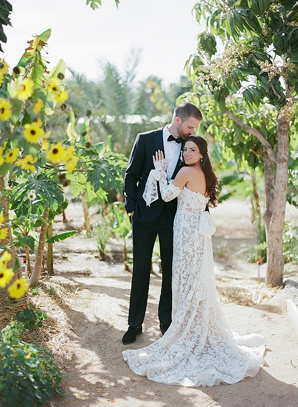 Couple portrait of groom kissing bride’s forehead as she wears an off-the-shoulder lace dress on a sunlit garden path with yellow flowers